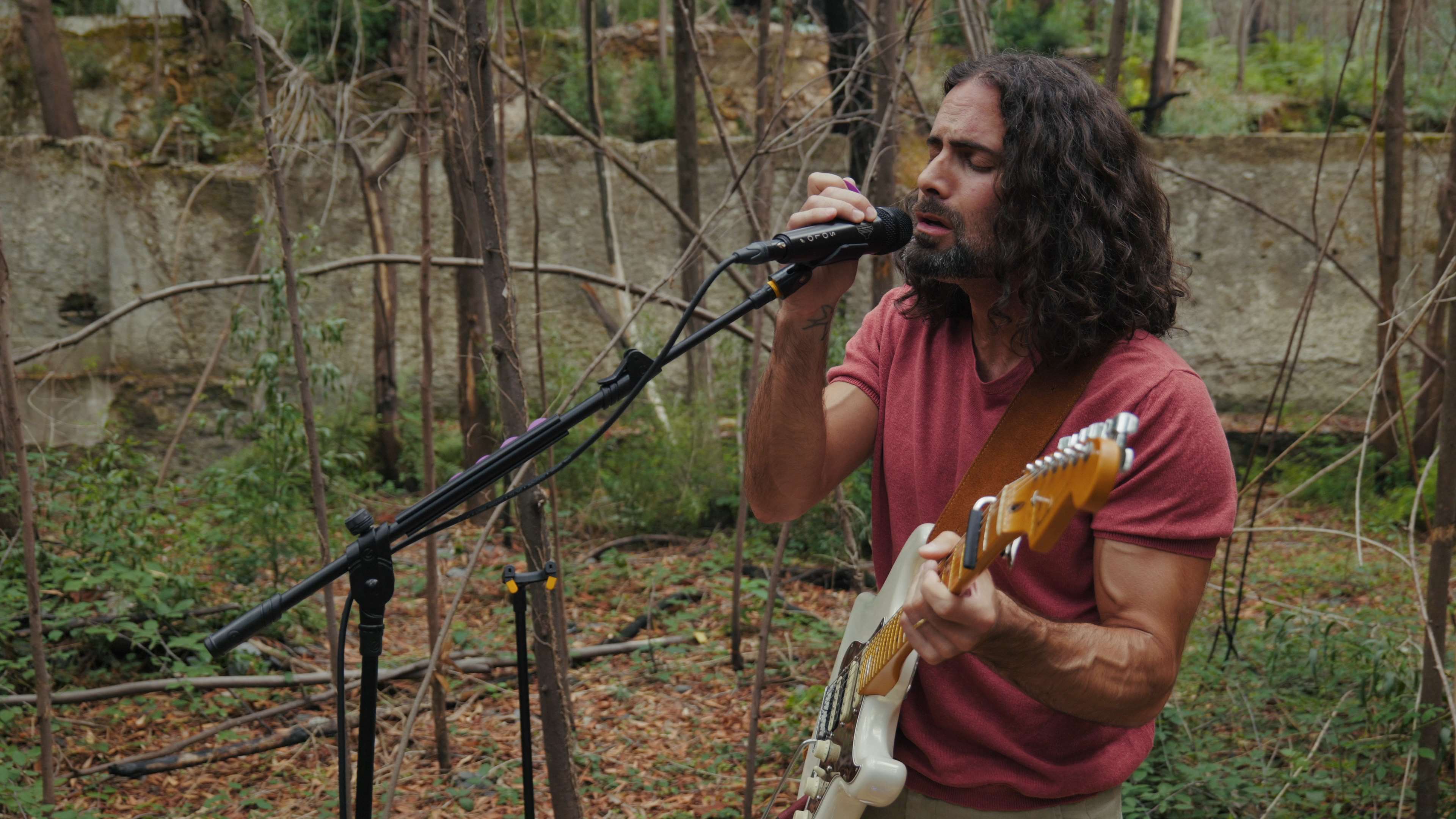 Homem com uma guitarra elétrica e a cantar para um microfone numas ruínas abandonadas repletas de vegetação